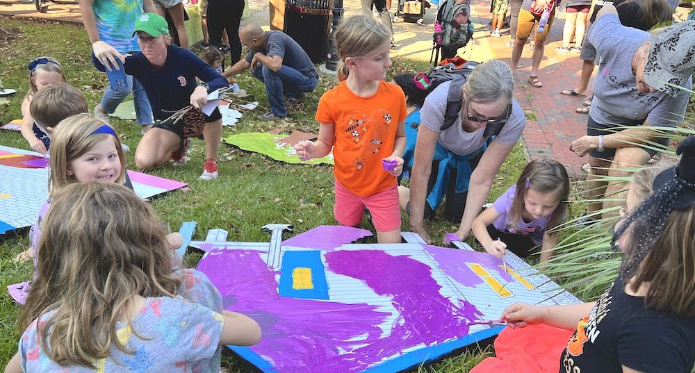 Kids paint colorful cardboard images of buildings on a grassy lawn.