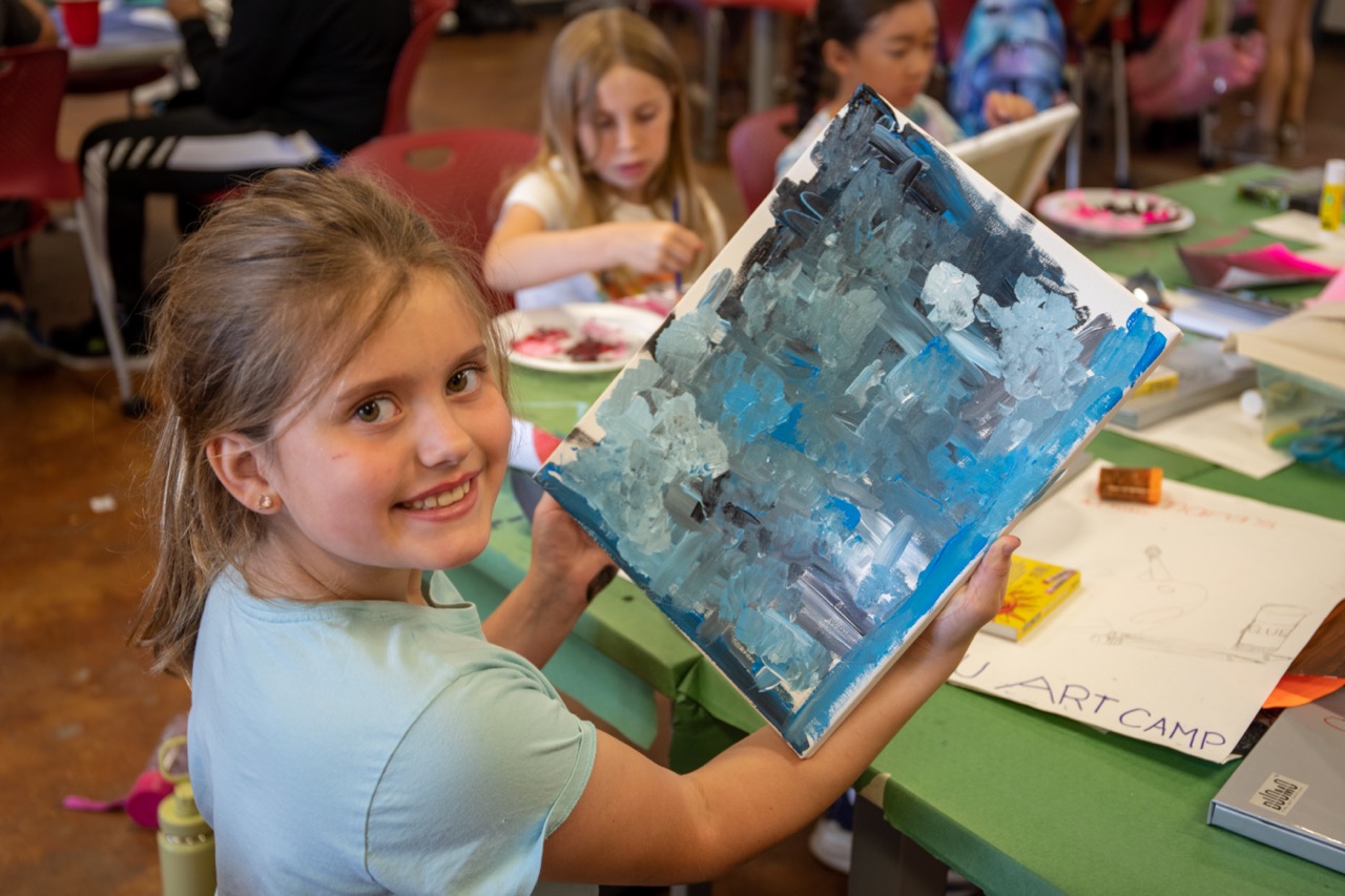 A little girl holds up a blue abstract painting that she is working on. She smiles brightly as she displays the work for the photo. 