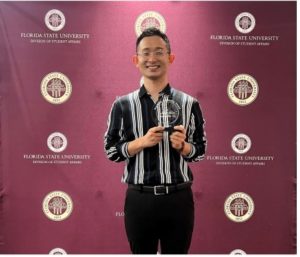 A man poses with an award in front of a garnet colored backdrop featuring the FSU seal. He is smiling and wearing a striped button up shirt and black pants. 