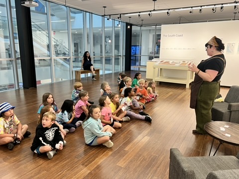 there is a woman standing in front of a group of children sitting on the floor