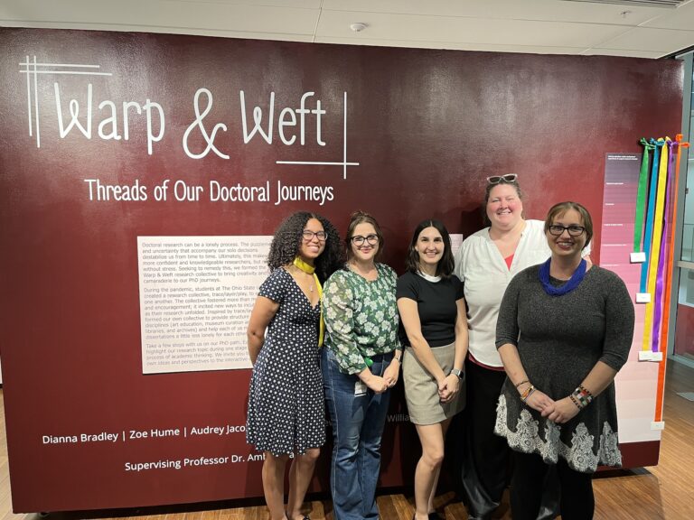 A group of students stand in front of a red wall with vinyl text on it that says "Warp & Weft"