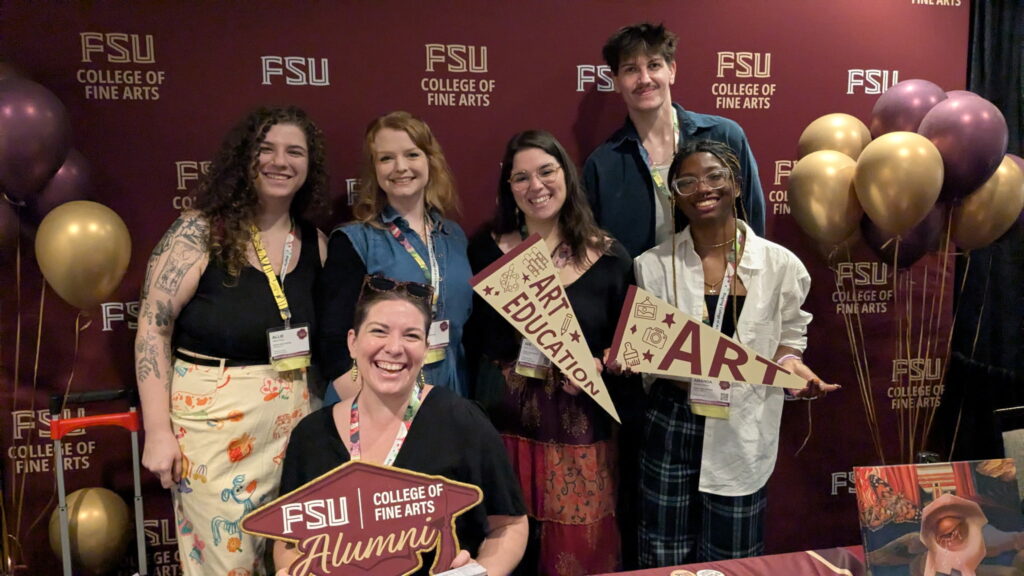 A group of smiling art education students hold up banners. and pose with balloons. 
