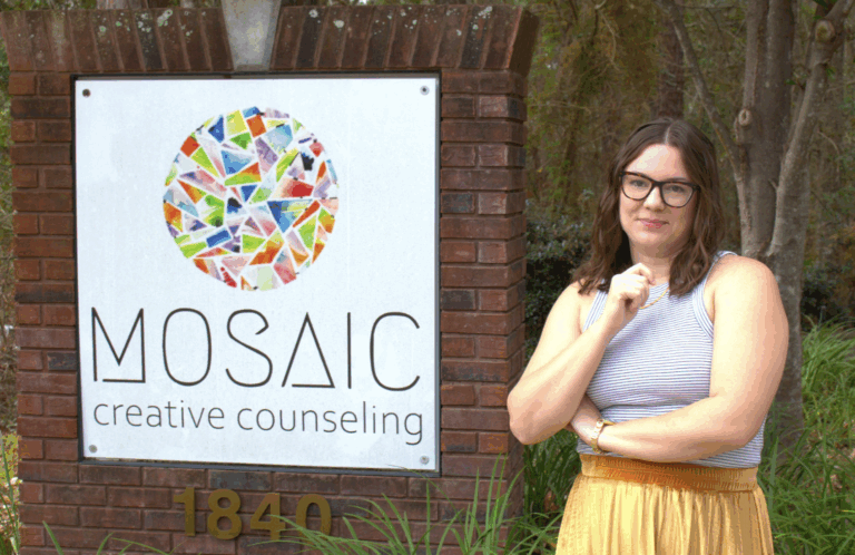 A woman standing next to a sign outside that says "Mosaic Creative Counseling" and has a big colorful circle on it.