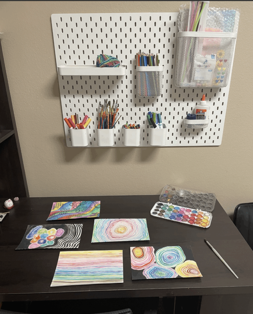 Colorful artwork is scattered on a table, a peg board with neatly organized art supplies hangs on the wall. 