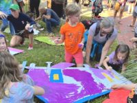 Kids paint colorful cardboard images of buildings on a grassy lawn.