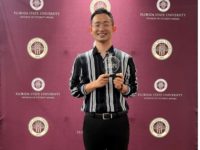 A man poses with an award in front of a garnet colored backdrop featuring the FSU seal. He is smiling and wearing a striped button up shirt and black pants.