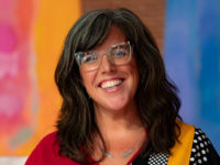 A woman wearing glasses is smiling in front of a colorful background